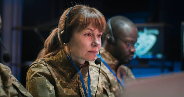 Multiethnic male and female military dispatchers in camouflage uniforms and headsets working at monitoring center at computers. Caucasian and African American men and woman in army office.