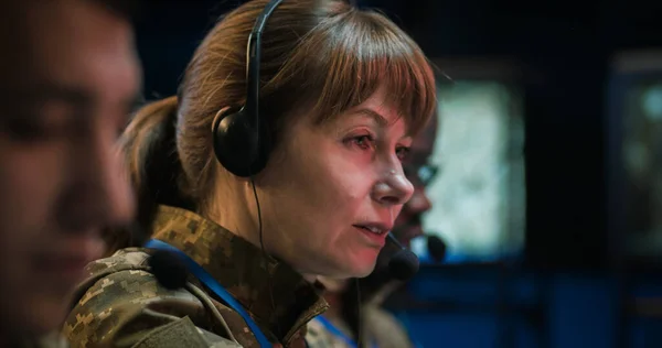 Caucasian military woman in headset sitting at computer and talking via videochat in controlling center of army. Female officer speaking via webcam in military office. Mixed-races dispatchers.