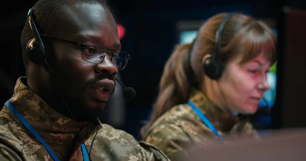 Close up of African American male officer veteran in headset working as dispatcher in monitoring room. Soldier answering calls on hot line of army controlling center. Caucasian woman on background.
