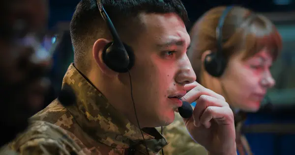 Close up of Caucasian male officer in headset working as dispatcher in monitoring room. Soldier answering calls on hot line of army controlling center. Multiethnic male and female co-workers beside.