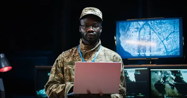 Portrait shot of African American handsome young man army general standing in monitoring room and using laptop computer. Soldier looking straight and smiling to camera.