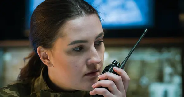 Close up of Caucasian young woman in army uniform sitting in monitoring room and talking, using walkie-talkie. Female soldier speaking in radio transmitter in troops controlling office.
