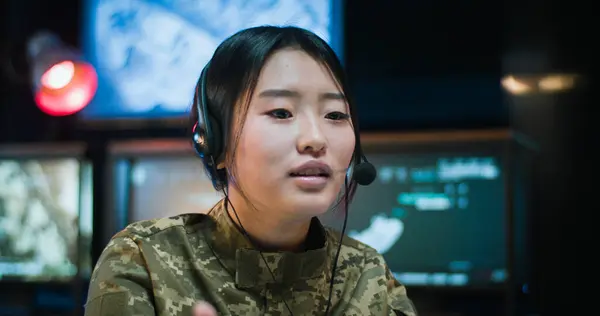 Close up of Asian young woman in camouflage clothes and headset sitting at computer screen in monitoring room in army and talking via webcam. Female soldier working in military data center.