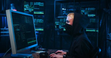 Yandan bakan, maskeli beyaz kadın hacker büyük ekranın önünde bilgisayarda çalışıyor ve karanlık odada klavyede daktilo kullanıyor..