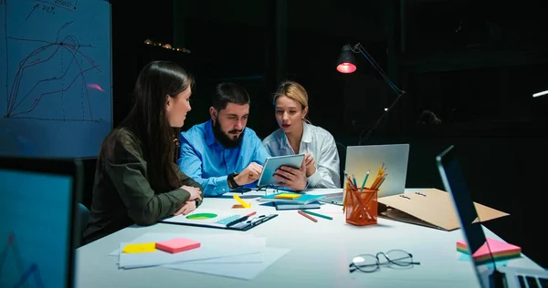 Caucasian man boss of company with beard sitting in office at nigh with two female co-workers and working at laptop computer. Business colleagues having meeting in evening and talking.