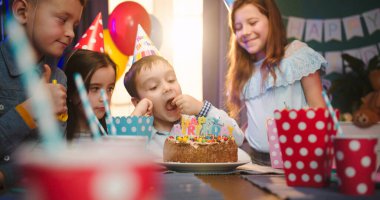 Sevimli küçük beyaz çocuk masanın üzerine eğilmiş ve doğum günü partisinde arkadaşlarının arasında pastaya bakmaktan sıkılmış..