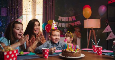 Doğum günü çocuğu ve annesi olan neşeli ve mutlu beyaz çocuklar ellerinde pastayla masada oturuyorlar ve arka plandaki renkli konfetilerde doğum günlerini kutluyorlar. Parti ev dekorasyonu.