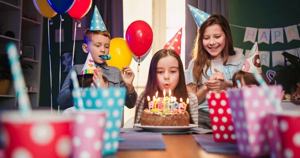 Happy little girl celebrating birthday with friends at home, blowing candles at cake , making wish while sitting at table in living room and children blowing in the party horns.