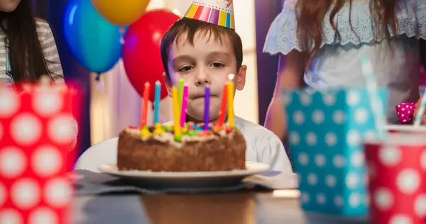 Sad little boy celebrating birthday with family at home, looking at cake with lit candles, making wish while sitting at table in living room in colorful conus