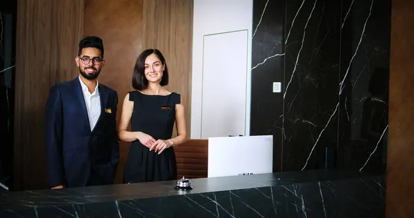 Portrait of polite and hospitable young man and woman working as hotel staff at reception. Handsome Indian male receptionist in suit and elegant female manager in dress looking at camera and smiling.