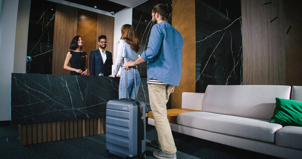 Young lovely couple of man and woman approaching hotel reception with male and female receptionists welcoming new guests. Two attractive people with luggage checking in.