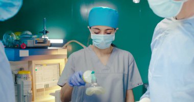 Özel kıyafetli sağlık görevlileri. Cerrahi odasındaki tıbbi ekipmanların arka planındaki doktorlar. Afrikalı Amerikalı asistan elinde pamuklu cımbız tutuyor. Stajyer hastalara tüp yutturuyor.