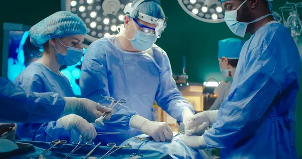 Diverse team of professional surgeons performs invasive surgery on patient in hospital. Nurse gives necessary tools to doctor. African-American assistant helps head surgeon apply sutures.