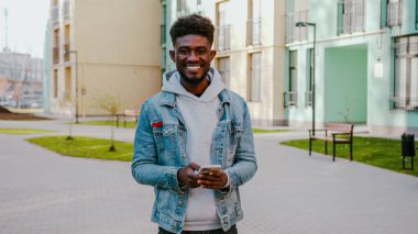 Elinde modern akıllı telefonuyla caddede duran gülümseyen Afro-Amerikan adam. Gündelik giyinen, internette sörf yapan ya da yalnız yürürken sohbet eden pozitif bir genç..