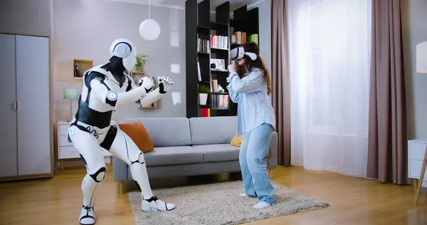 Side view of human like robot fist bumping with attractive woman in VR goggles in living room. Concept of people, innovation and artificial intelligence.