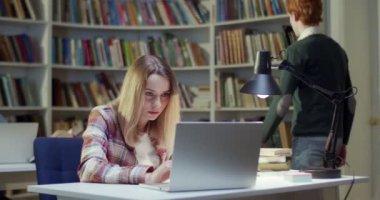 Kütüphanede dizüstü bilgisayar okuyan ve internetten bilgi arayan sarı saçlı beyaz bir kız öğrenci. Sınavlara hazırlanıyorum. öğrenim kavramı.