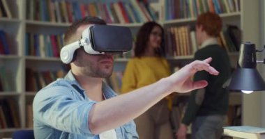 VR kulaklıklı genç beyaz adam kütüphane alanında oturuyor, ellerini havada kaydırıyor ve hareket ettiriyor ve sanal gerçeklik deneyimi yaşıyor. Metaverse oyun. Oyun oynamak ya da sanal ortamda çalışmak. Kapat..