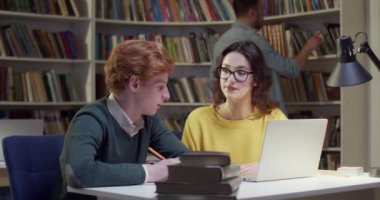 Beyaz bir kadın kütüphanede sınava hazırlanırken erkek öğrenciye yardım ediyor. Açıklama dersi materyali. Arkadaşlar bibliyotheca 'da dizüstü bilgisayarla birlikte öğreniyor. Öğrenciler konuşuyor ve çalışıyor..