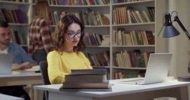 Gözlüklü genç bayan öğrenci dizüstü bilgisayarda kütüphane okuyor ve sıkılıyor. Kitap konseptinde sıkıcı bir çalışma. Kız öğrenme ve sınava hazırlanma.