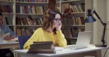Gözlüklü güzel Cacasian genç kız öğrenci dizüstü bilgisayarda kütüphane okuyor ve sıkılıyor. Kitap konseptinde sıkıcı bir çalışma. Kız öğrenme ve sınava hazırlanma.