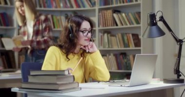 Kulaklıklı, gözlüklü güzel bir kadın kütüphanede dizüstü bilgisayarla konuşuyor, yazışıyor ve sohbet ediyor. Konuşan, notlar alan ve webcam üzerinden görüntülü sohbet yapan bir kadın. Görüntülü arama. Not bilgileri.