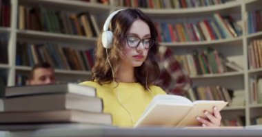 Gözlüklü, kulaklıklı, kitap okuyan ve kütüphanede müzik dinleyen güzel beyaz bir kadın. Bibliyotheca 'da eğitici bir kavram. Bayan öğrenci şarkı dinleyip ders çalışıyor..