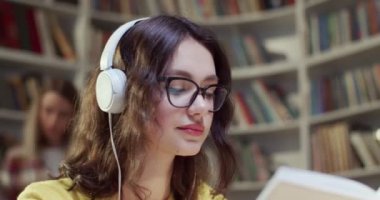 Gözlüklü, kulaklıklı, kitap okuyan ve kütüphanede müzik dinleyen güzel beyaz bir kadın. Bibliyotheca 'da eğitici bir kavram. Bayan öğrenci şarkı dinleyip ders çalışıyor..