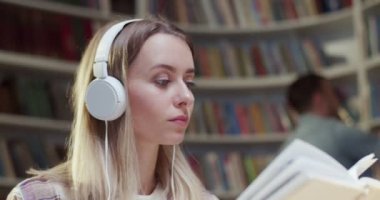 Kulaklıklı, kitap okuyan ve kütüphanede müzik dinleyen güzel beyaz bir kadın. Bibliyotheca 'da eğitici bir kavram. Bayan öğrenci şarkı dinleyip ders çalışıyor..
