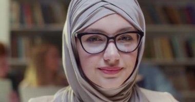 Kütüphanede oturmuş kameraya gülümseyen tesettürlü, tesettürlü, tesettürlü, genç ve güzel bir Arap kadını yakından çek. Bibliyotheca 'daki Arap türbanlı güzel kız öğrenci. Portre.