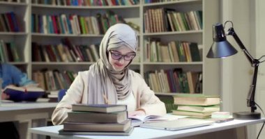 Genç tesettürlü, tesettürlü, gözlüklü, kitap okuyan ve sayfaları işaretleyen Müslüman bir kız öğrenci. Arap kadın başörtüsü takıyor ders kitabı okuyor ve işaretliyor..