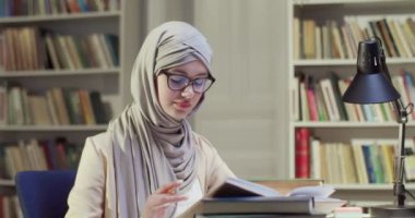 Genç tesettürlü, tesettürlü ve gözlüklü Müslüman kız öğrenci kütüphanede oturmuş, notlar alıyor ve ders kitabı okuyor. Arap başörtüsü takan kadın kitaptan öğreniyor ve not alıyor. Bibliotheca çalışma konsepti.