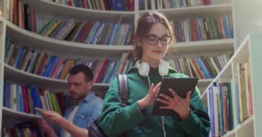 Kafkasyalı, gözlüklü genç kız öğrenci kütüphanede duruyor, tablet cihazı kullanıyor ve ekranda tıklıyor. Güzel bir kadın internette bilgi almak için mesajlaşıyor ve internette sörf yapıyor..