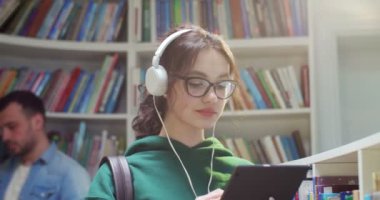 Gözlüklü ve kulaklıklı genç kız öğrencileri tablet cihazı kullanarak ve kütüphanede ekrana vurarak kapatın. Bilgi almak için internette sörf yapan şık bir kadın..