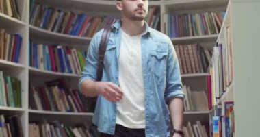Sırt çantalı bir erkek öğrenci kütüphanede yürüyor, okumak için ders kitabını seçiyor. Raftan kitap çıkarıyorum. Edebiyat konsepti. Bibliotheca 'ya okumaya gelen adam..