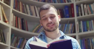 Beyaz tenli yakışıklı erkek öğrencinin sayfaları çevirmesi, kitap okuması ve kütüphanede bilgi araması. Kitaplıkta kitabı olan adam neşeyle gülümsüyor. Portre çekimi.