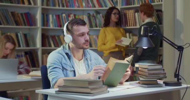 白种人年轻男生在耳机翻页的旧教科书和学习 在图书馆听音乐 在书目中读书的家伙 教育概念 — 图库视频影像
