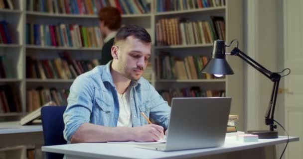 在图书馆的笔记本电脑前学习的白人年轻人 男人在桌上做笔记 用铅笔在笔记本上写字的人 在书目中工作A 学习和工作概念 搜寻资料 — 图库视频影像