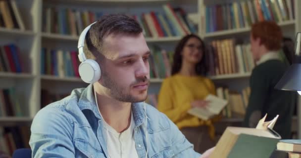 接近白种人的年轻男生在耳机翻页的旧教科书和学习 在图书馆听音乐 在书目中读书的家伙 教育概念 — 图库视频影像