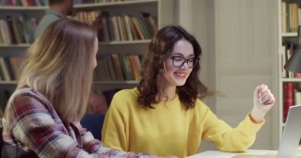 两个快乐的白种人漂亮的女人坐在桌旁 在图书馆学习时 害怕地谈笑着 在书目中快乐地聊天和微笑的女人 共同学习概念 — 图库视频影像