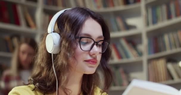 メガネとヘッドフォンで美しい若い白人女性が本を読み 図書館で音楽を聴いている イスラエルにおける教育の概念 女性学生が歌を聴き 勉強する — ストック動画
