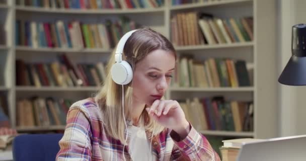 漂亮的金发白人女人 戴着耳机 在笔记本电脑上工作 在图书馆听音乐 书目中的教育概念A 女学生听歌曲 在电脑上敲键盘 — 图库视频影像