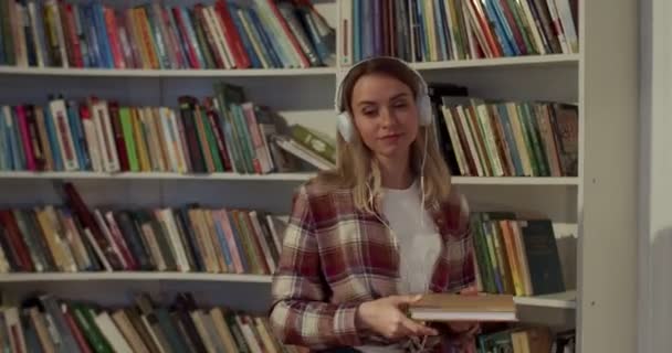 漂亮的白人女学生戴着耳机 在图书馆里挑选课本 然后坐在桌旁学习 金发女人从书目书架上拿出一本书阅读 — 图库视频影像