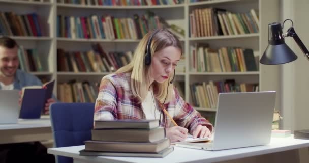 Piękna Blondynka Słuchawkach Mówiąca Pisząca Nagrywająca Laptopie Bibliotece Mówi Kobieta — Wideo stockowe