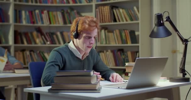 Biały Rudy Mężczyzna Słuchawkach Mówiący Piszący Nagrywający Laptopie Bibliotece Mówi — Wideo stockowe