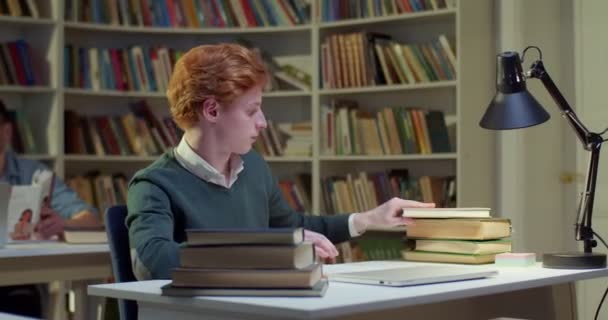 年轻的白种人男生选择在图书馆的餐桌边看书 并打开书柜看书 红头发的男人拿着课本去读 书目中的学习概念 考试的准备工作 — 图库视频影像