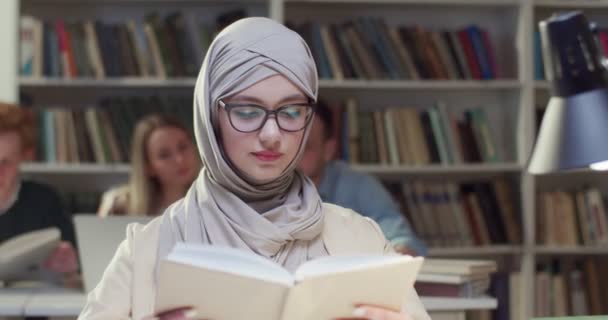 一名身穿头巾和眼镜的年轻阿拉伯穆斯林女子在图书馆看书 戴头巾的阿拉伯漂亮女生拿着课本 教育概念 学习和学习能力 — 图库视频影像