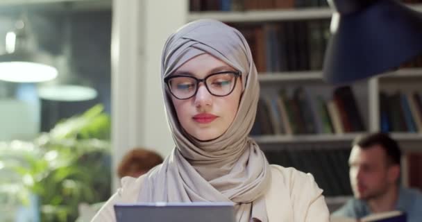接近年轻的阿拉伯穆斯林妇女在头巾和眼镜工作的平板在图书馆 阿拉伯漂亮的女生头戴头巾 手里拿着电脑小玩意 教育概念 在东方的研究 — 图库视频影像