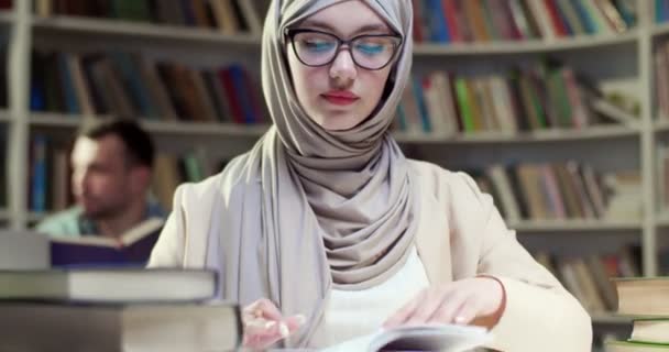 年轻漂亮的穆斯林女学生 头戴头巾 戴着眼镜 一边看书一边翻页 阿拉伯妇女在图书馆学习带课本和标识的头巾 — 图库视频影像