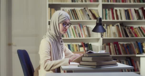 关于身穿头巾和眼镜的年轻穆斯林女学生坐在图书馆桌子边 记录笔记和阅读课本的侧视图 穿着阿拉伯头巾的女人注意并从书本中学习 学习概念 — 图库视频影像