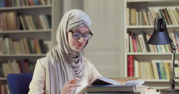 身穿头巾和眼镜的年轻穆斯林女学生坐在图书馆的桌旁 记录笔记和阅读课本 穿着阿拉伯头巾的女人注意并从书本中学习 参考书目研究概念 — 图库视频影像